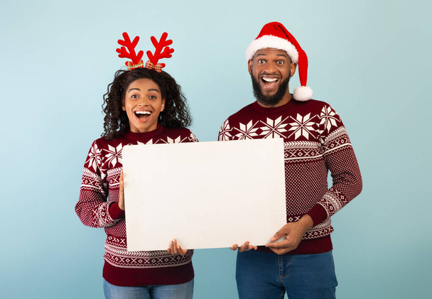 Χριστουγεννιάτικη διαφήμιση. Ευτυχισμένοι μαύροι σύζυγοι κρατώντας άδεια αφίσα, στέκεται πάνω από το μπλε φόντο, πανό - Φωτογραφία, εικόνα
