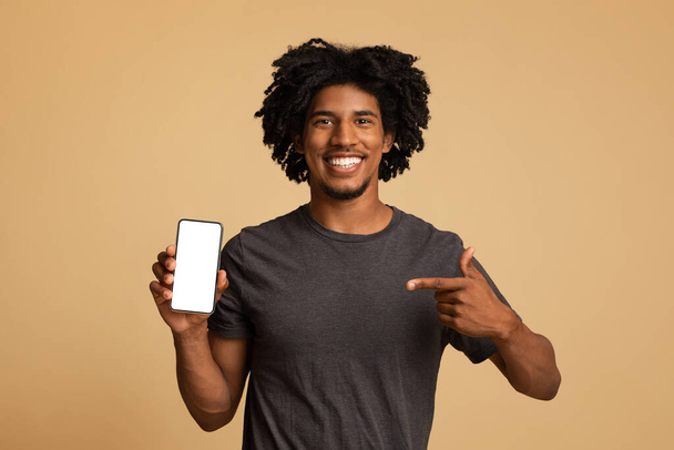 Fröhlicher schwarzer Kerl zeigt auf Smartphone mit weißem Bildschirm in der Hand - Foto, Bild