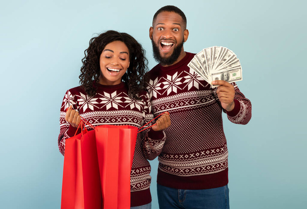 Χριστουγεννιάτικα ψώνια. Ευτυχισμένο ζευγάρι Αφροαμερικανών με Χριστουγεννιάτικα πουλόβερ που κρατάνε τσάντες για ψώνια και πολλά λεφτά. - Φωτογραφία, εικόνα