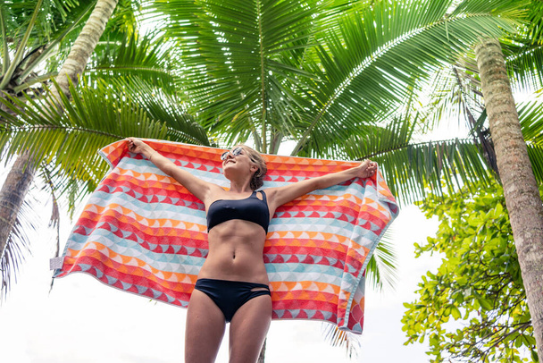 Blond vrouwtje in zonnebril en bikini houdt een kleurrijke handdoek voor een palmboom. - Foto, afbeelding