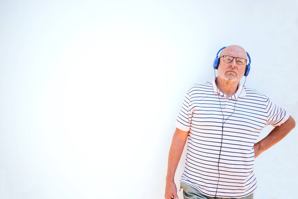 Homme âgé chauve détendu appuyé sur le mur tout en écoutant de la musique avec des écouteurs bleus sur un fond blanc - Photo, image