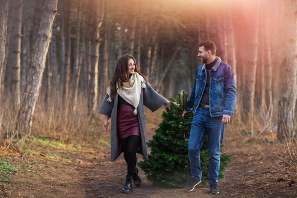 Happy man and woman in warm clothes carrying Christmas fir tree together walking on path in woods  - Valokuva, kuva
