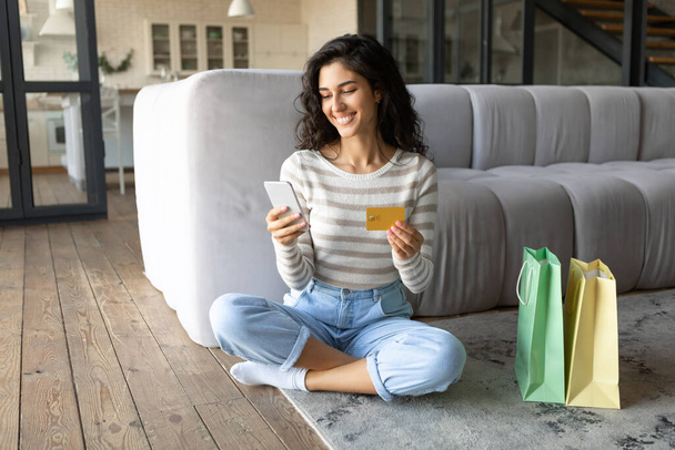 Joyeuse jeune femme utilisant une carte de crédit et un smartphone pour faire du shopping dans une boutique en ligne, assise sur le sol avec des sacs à provisions - Photo, image