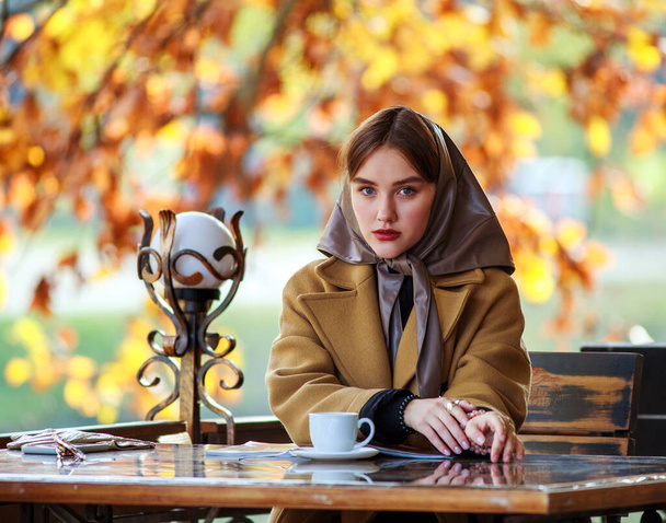 Luxurious young woman in retro fashion style, in a beige coat and scarf on her head, in a cafe on a wooden playground in autumn - Foto, Imagen