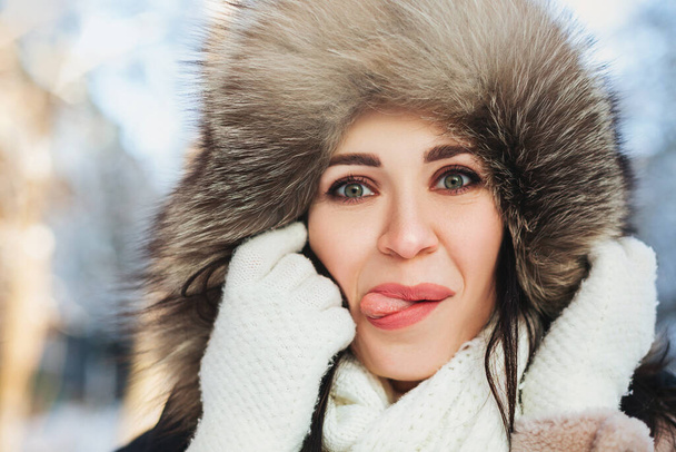 Engraçado jovem mulher no capuz peludo olhando para a câmera e mostrando a língua no fundo borrado da rua no dia de inverno - Foto, Imagem