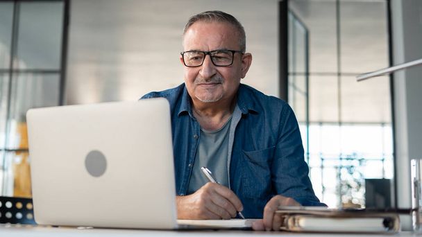 Hombre mayor mirando el ordenador portátil y tomando notas - Foto, imagen