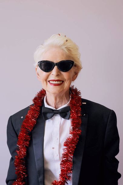 Piękny stylowy dojrzały starszy kobieta w okularach przeciwsłonecznych i smokingu świętuje Nowy Rok. Zabawa, impreza, styl, koncepcja uroczystości - Zdjęcie, obraz