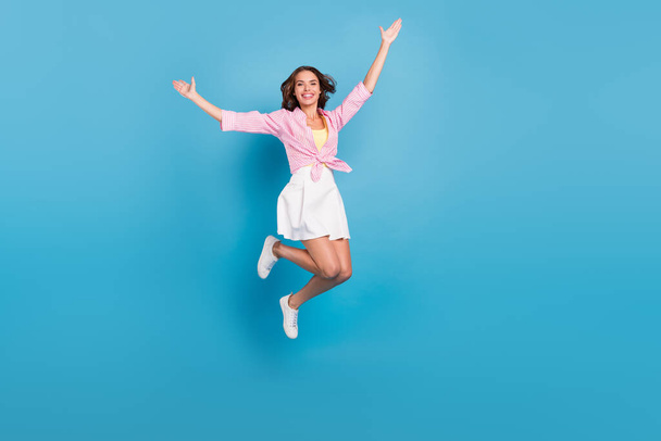 Photo of adorable shiny young woman dressed white skirt smiling rising hands arms jumping high isolated blue color background - Fotoğraf, Görsel