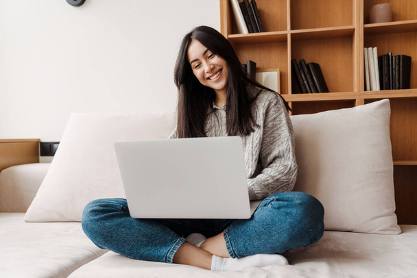 Glückliche hübsche Frau benutzt Laptop-Computer, während sie es sich zu Hause auf der Couch gemütlich macht - Foto, Bild