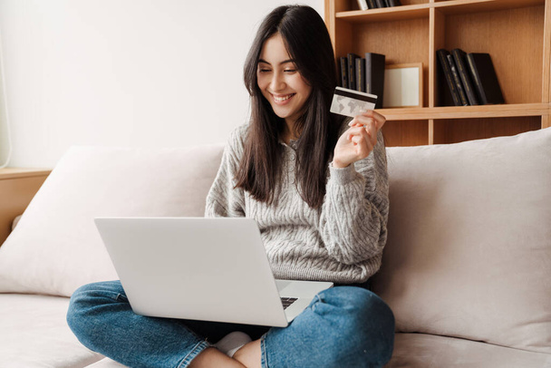 Щаслива молода приваблива жінка купує онлайн через ноутбук, використовуючи кредитну картку вдома
 - Фото, зображення