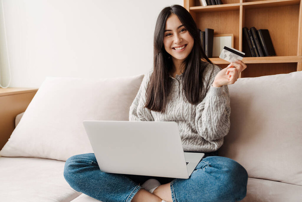 Happy young attractive woman shopping online via laptop computer, using credit card at home - Foto, Imagen