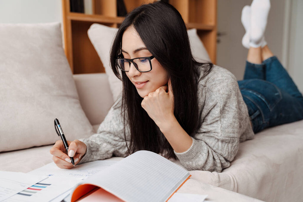 Felice asiatico studente ragazza fare compiti mentre sdraiato su divano a casa - Foto, immagini