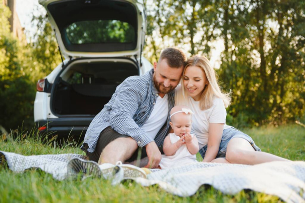 Young family three people in white clothes have picnic. Beautiful parents and daughter travel by car during summer vacation. - Zdjęcie, obraz