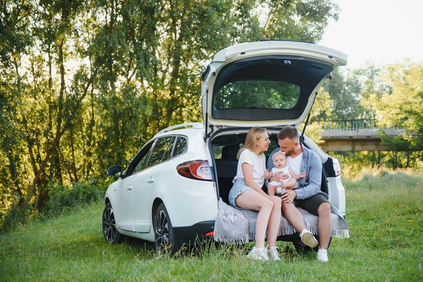 Pretty young married couple and their daughter are resting in the nature. The woman and girl are sitting on open car boot. The man is standing near them. They are smiling - Foto, Bild