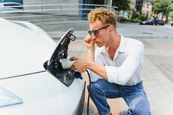jonge knappe man met oplaadkabel op elektrisch laadstation punt staan in de buurt van zijn nieuwe auto, op zoek tevreden. - Foto, afbeelding