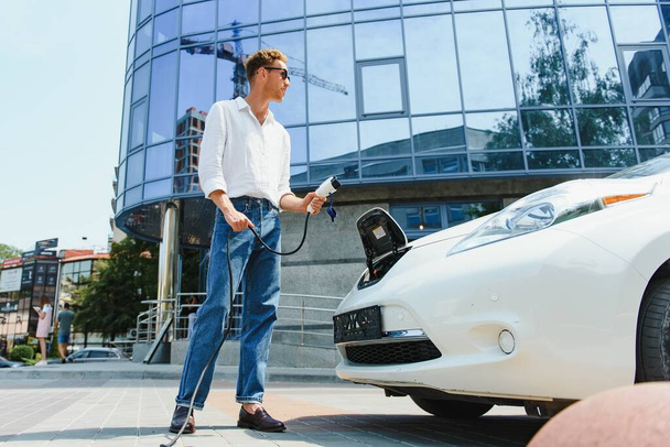 Man Holding Power Charging Cable For Electric Car In Outdoor Car Park. And he s going to connect the car to the charging station in the parking lot near the shopping center - Fotoğraf, Görsel