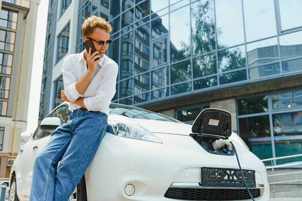 Όμορφος άνθρωπος που χρησιμοποιεί το τηλέφωνο, ενώ το αυτοκίνητο χρεώνεται - Φωτογραφία, εικόνα