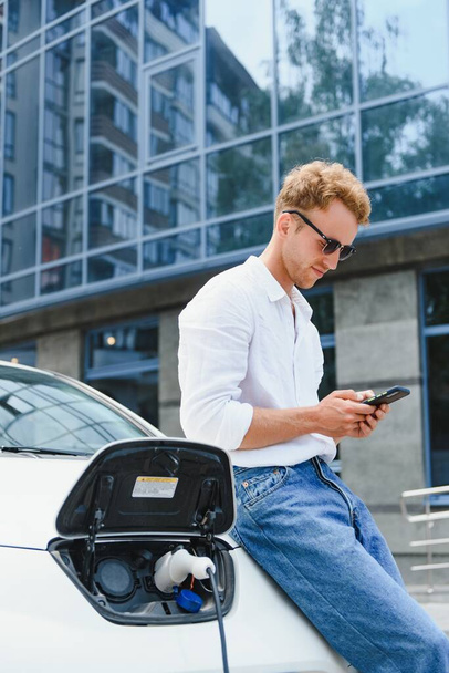 Man with smartphone after finishing charging of battery. Male near electric vehicle - Fotoğraf, Görsel