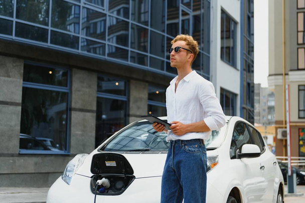 portrait of young handsome man in casual wear, standing at the charging station. Eco electric car concept - Fotografie, Obrázek