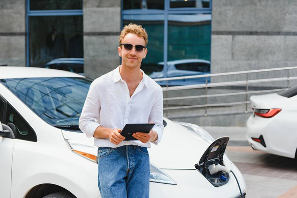 Business man standing near charging electric car and using tablet in the street. - Foto, Imagem