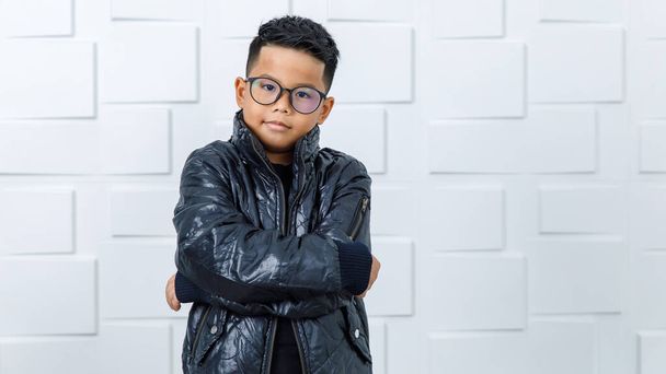 Портрет молодого красивого азиатского мальчика в очках и черной куртке, стоящего у белой стены, улыбающегося, сжимающего правый кулак и поднимающего локоть так же уверенно и уверенно в своем решении - Фото, изображение