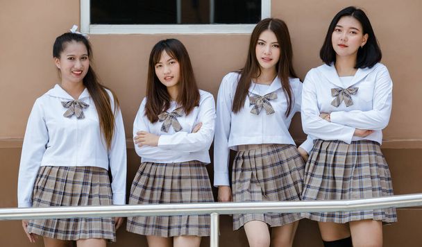 Grupa czterech młodych atrakcyjnych azjatyckich uczennic liceum w brązowym kratowym mundurku szkolnym stojących razem w kampusie szkolnym uśmiechając się. Koncepcja edukacji i życia uczniów szkół średnich. - Zdjęcie, obraz