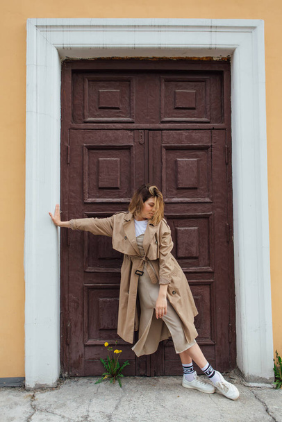 Young smiling millennial woman with wild hair dressed in an autumn coat posing near the door of an old building. - Foto, Imagem