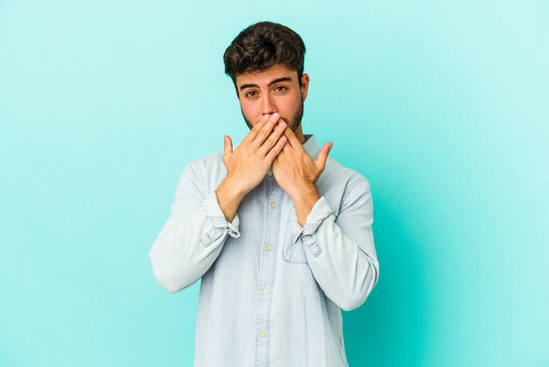 Young caucasian man isolated on blue background shocked covering mouth with hands. - Zdjęcie, obraz