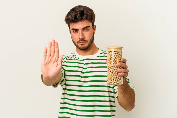 Νεαρός καυκάσιος άνδρας που κρατά το βάζο με τα ρεβίθια απομονωμένο σε λευκό φόντο στέκεται με τεντωμένο χέρι που δείχνει στοπ, εμποδίζοντάς σας. - Φωτογραφία, εικόνα