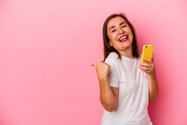 Середньовічна біла жінка тримає мобільний телефон ізольовано на рожевих точках тла з пальцем на великий палець, сміється і безтурботний
. - Фото, зображення