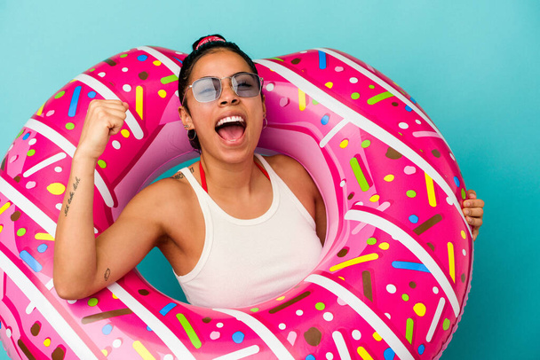 Mavi arka planda izole edilmiş şişme donut tutan genç Latin kadın zafer kazandıktan sonra yumruğunu kaldırıyor, kazanan konsept.. - Fotoğraf, Görsel