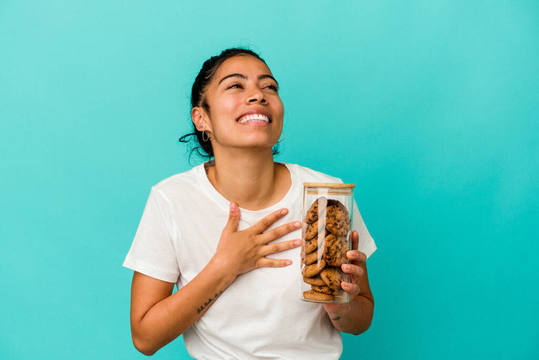 Νεαρή Λατίνα γυναίκα κρατώντας ένα βάζο με μπισκότα απομονωμένο σε μπλε φόντο γελάει δυνατά κρατώντας το χέρι στο στήθος. - Φωτογραφία, εικόνα