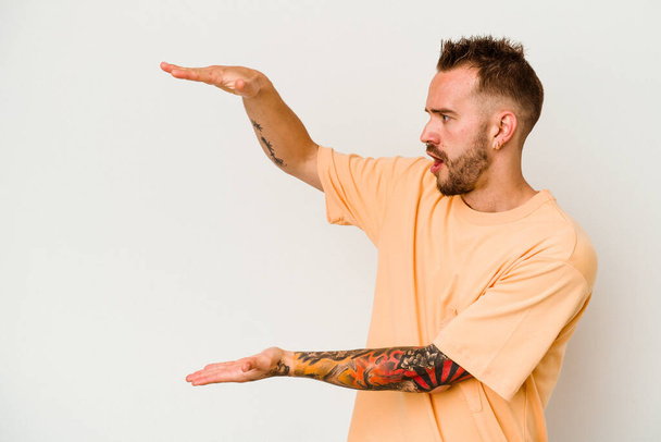 Young tattooed caucasian man isolated on white background shocked and amazed holding a copy space between hands. - Фото, изображение
