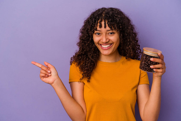 Joven mujer de raza mixta sosteniendo una botella de granos de café aislado sobre fondo púrpura sonriendo y señalando a un lado, mostrando algo en espacio en blanco. - Foto, Imagen