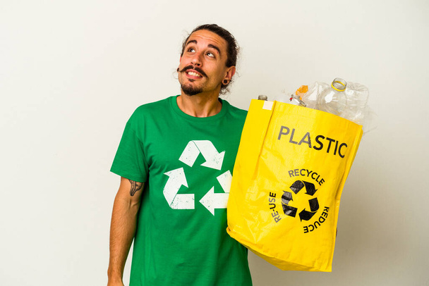 Junge kaukasische Mann mit langen Haaren Recycling-Kunststoff isoliert auf weißem Hintergrund träumen vom Erreichen von Zielen und Zwecken - Foto, Bild