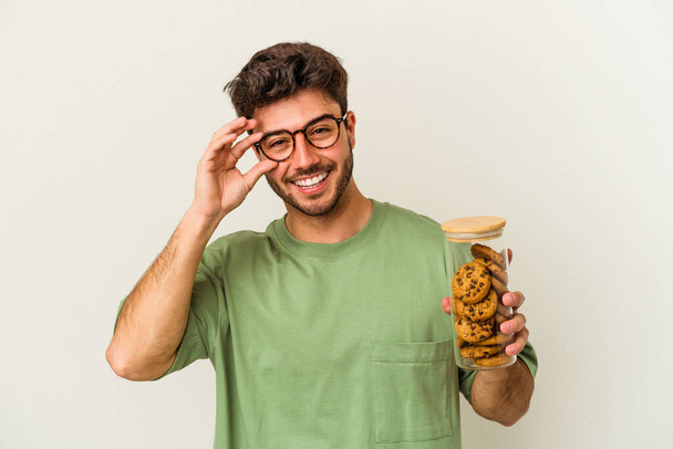 Young caucasian man holding cookies jar isolated on white background excited keeping ok gesture on eye. - Photo, Image