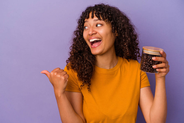Młoda mieszanka rasy kobieta trzyma butelkę ziarna kawy izolowane na fioletowym tle punktów kciukiem daleko, śmiejąc się i beztroski. - Zdjęcie, obraz