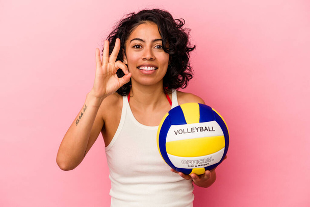 Giovane donna latina in possesso di una pallavolo isolato su sfondo rosa allegro e fiducioso mostrando gesto ok. - Foto, immagini