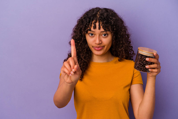 Joven mujer de raza mixta sosteniendo una botella de granos de café aislado sobre fondo púrpura que muestra el número uno con el dedo. - Foto, Imagen