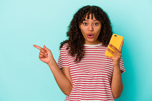 Junge Frau mit gemischter Rasse hält ein Handy isoliert auf blauem Hintergrund und zeigt zur Seite - Foto, Bild