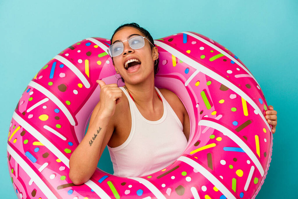 Junge lateinische Frau hält einen aufblasbaren Donut isoliert auf blauem Hintergrund mit dem Daumenfinger weg, lacht und ist unbeschwert. - Foto, Bild