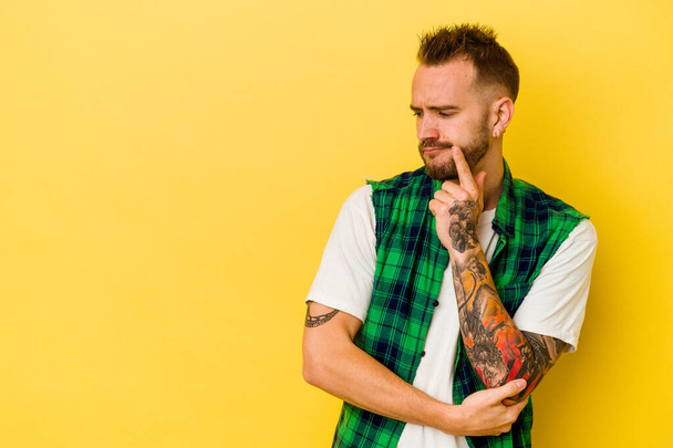 Young tattooed caucasian man isolated on yellow background looking sideways with doubtful and skeptical expression. - Photo, Image