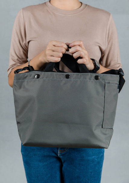Closeup studio shot of female model in brown long sleeve shirt and jeans stand holding handle of fashionable trendy urban big blue unisex handbag with long shoulder strap in front of grey background. - Fotografie, Obrázek