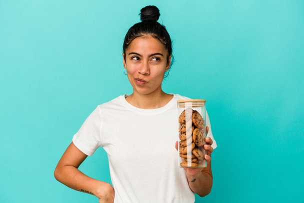 Mujer latina joven sosteniendo un tarro de galletas aislado sobre fondo azul confundido, se siente dudoso e inseguro. - Foto, imagen