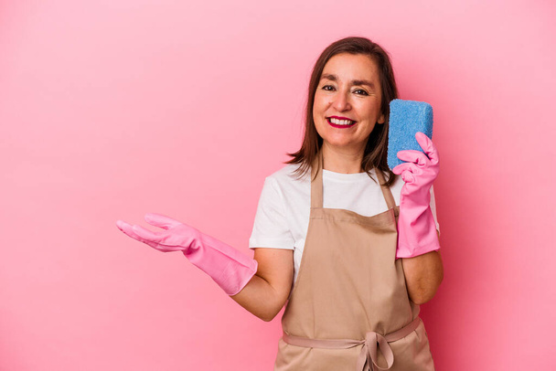 Mulher caucasiana de meia idade limpando casa isolada em fundo rosa mostrando um espaço de cópia em uma palma e segurando outra mão na cintura. - Foto, Imagem