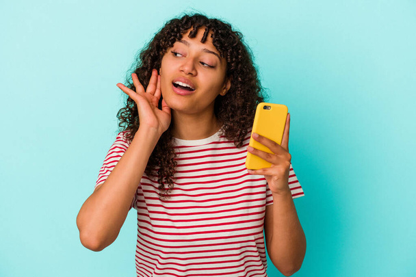 Fiatal vegyes fajú nő kezében egy mobiltelefon elszigetelt kék háttér próbál hallgatni egy pletyka. - Fotó, kép