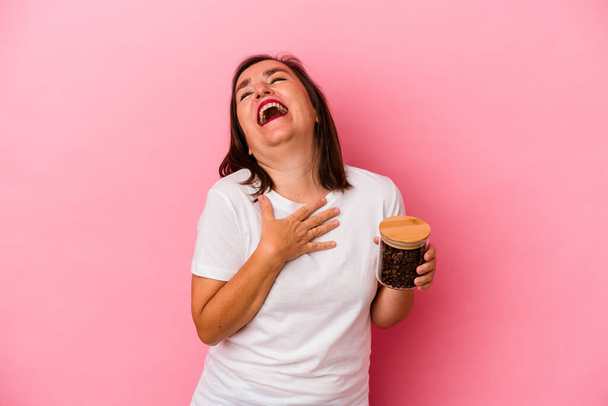 Середньовічна біла жінка тримає кавову банку ізольовану на рожевому фоні, голосно сміється, тримаючи руку на грудях
. - Фото, зображення