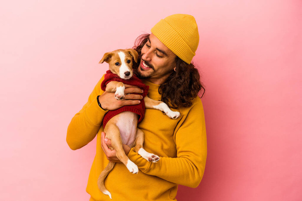 jonge Kaukasische man het houden van zijn puppy geïsoleerd op roze achtergrond - Foto, afbeelding