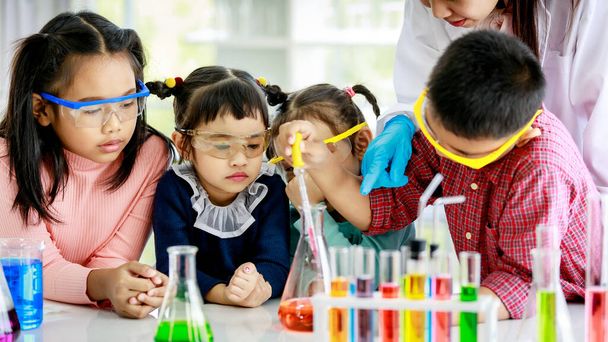 Asian little boy drop reagent sample by glass dropper into colorful solution flask while young girls wears safety glasses and teacher in white lab coat look at him do experiment in school laboratory. - Photo, Image