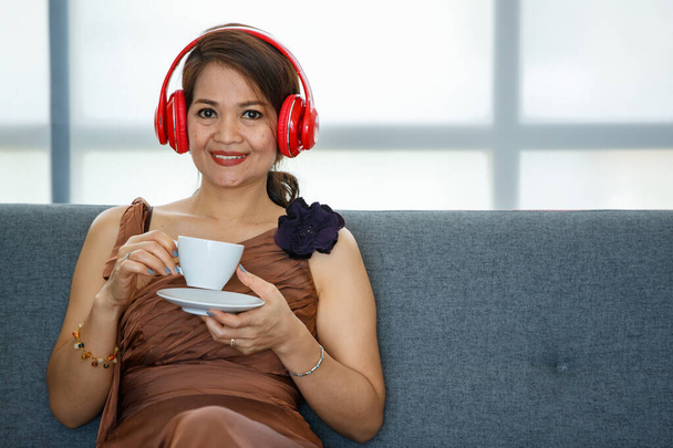 50s πλούσια ασιατική αριστοκρατική πολυτέλεια γυναίκα, κομψό καλοντυμένο, κάθεται στο άνετο σαλόνι, βάζοντας ακουστικά και ακούγοντας μουσική, ενώ κρατώντας ένα φλιτζάνι καφέ και πίνοντας με την ευτυχία - Φωτογραφία, εικόνα